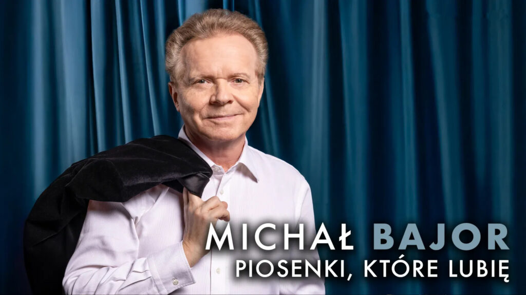 Portret uśmiechniętego Michała Bajora w białej koszuli z marynarką zarzuconą na ramię, na tle niebieskiej zasłony