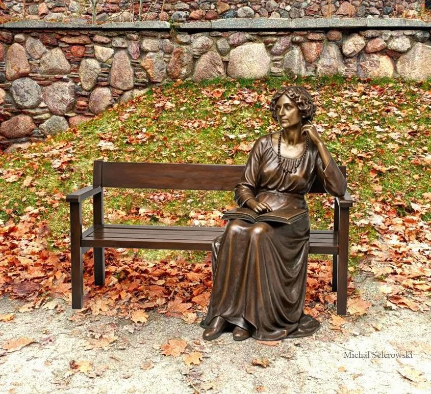 Brązowa rzeźba kobiety na ławce trzymającej książkę.