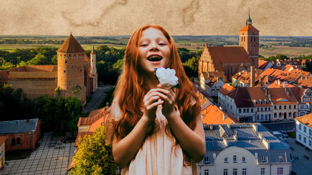 Uśmiechnięta dziewczynka z rudymi włosami trzyma kwiat na tle miasta.